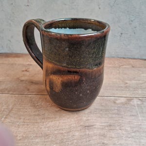 May include: A ceramic mug featuring a dark brown and olive green glazed exterior with a smooth, rounded body and a sturdy handle. The interior of the mug is finished with a contrasting light cream or off-white glaze, visible at the top rim.