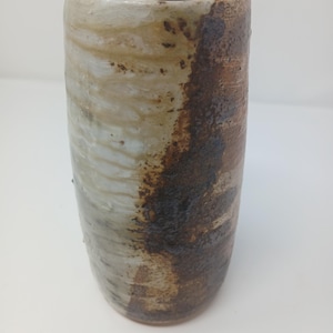 Könnte beinhalten: Ein handgefertigter Keramikbecher mit zylindrischer Form. Die Außenseite zeigt eine Mischung aus Farben, darunter Weiß, Beige und Dunkelbraun, wodurch ein rustikales, strukturiertes Aussehen entsteht. Der Becher ist für Getränke geeignet.