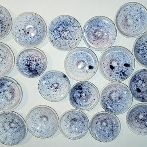 Könnte beinhalten: Eine Sammlung von 19 kleinen, weißen Keramikschalen mit einem gesprenkelten blauen und violetten Überzug. Die Schalen sind in einem Halbkreis auf einer weißen Oberfläche angeordnet.