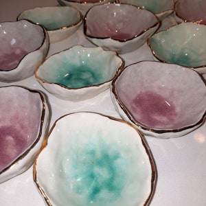 May include: A collection of small, decorative bowls with a white base and a gradient of pink and green glaze. The bowls have a gold rim and an irregular, organic shape.