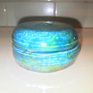 May include: A small, round, blue and green ceramic trinket box with a lid. The box has a swirling pattern and is made of glazed pottery.
