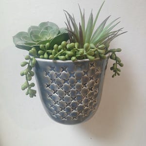 Puede incluir: Una maceta de pared de cerámica azul con un patrón de cruces, llena de suculentas artificiales variadas. El arreglo incluye una suculenta grande y redonda, varias plantas puntiagudas y plantas verdes con forma de cuentas.