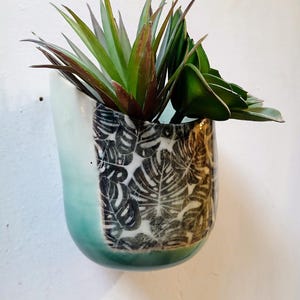 May include: A ceramic wall planter with a teal and white glaze, featuring a black and white leaf pattern. The planter holds a variety of green and burgundy artificial plants, creating a decorative display. The planter is mounted on a white wall.