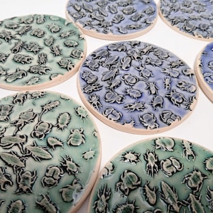 Può includere: Una collezione di sottobicchieri in ceramica con un motivo a rilievo di insetti. I sottobicchieri sono in tonalità di blu e verde, con un bordo color crema. I dettagli degli insetti sono in una tonalità più scura.