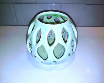 Puede incluir: Una esfera de cerámica verde claro con un diseño de celosía. La esfera tiene cortes ovalados y una superficie texturizada. El interior es de un tono verde más oscuro. Un elemento decorativo para el hogar.