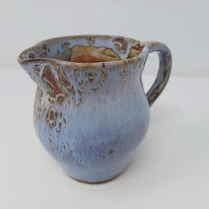 May include: A handmade ceramic pitcher with a blue and brown glaze. The pitcher has a rounded body, a handle, and a spout for pouring. The glaze has a textured appearance, adding to its unique, artisanal charm. This decorative pitcher is perfect for serving beverages.