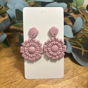Puede incluir: Un par de pendientes de color rosa empolvado con un diseño de flor circular y recortes de pétalos intrincados. Cada pendiente está unido a un pequeño poste en forma de diamante. Los pendientes se presentan en una tarjeta blanca rectangular.