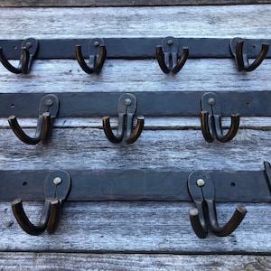 May include: Three wrought iron coat racks with multiple hooks. Each rack is a dark, matte black with a textured surface. The hooks are a darker, bronze color. The racks are mounted on a weathered wooden surface.