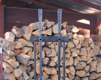 May include: A wrought iron fireplace tool set with a shovel, poker, and brush. The tools are displayed on a stand and are set against a backdrop of stacked firewood. The tools are made of dark metal.