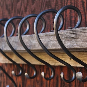 May include: A row of black metal hooks with a curved design, mounted on a weathered wooden plank. The hooks are attached to a dark, reddish-brown wooden surface, creating a rustic aesthetic.