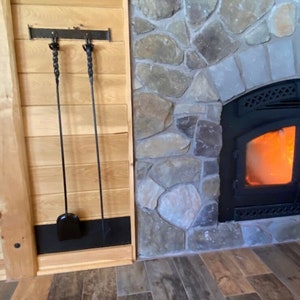 K&ouml;nnte beinhalten: Ein Kamin mit Steinfassung und brennendem Feuer. Ein Satz schwarzer Kaminwerkzeuge h&auml;ngt an der Holzverkleidung auf der linken Seite. Die Werkzeuge umfassen eine Schaufel und einen Sch&uuml;rhaken.