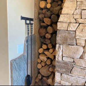 K&ouml;nnte beinhalten: Ein schwarzes Schmiedeeisen-Kaminwerkzeug-Set, das an einer Wand h&auml;ngt. Das Set enth&auml;lt einen Sch&uuml;rhaken und eine Schaufel. Die Werkzeuge befinden sich neben einem Stapel Brennholz und einem Steinkamin.
