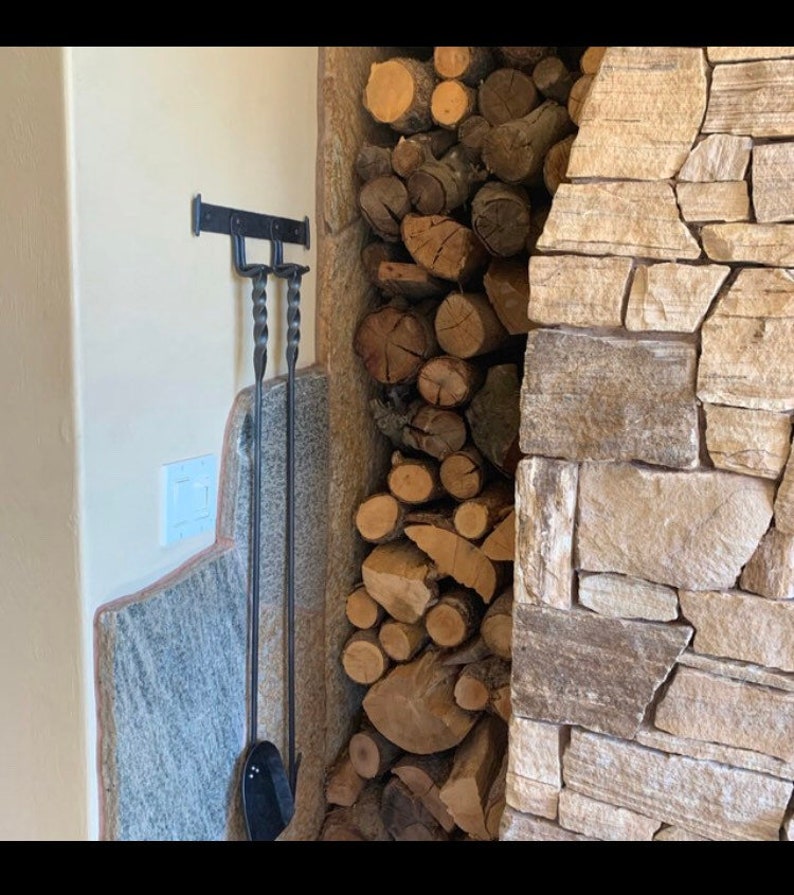 K&ouml;nnte beinhalten: Ein schwarzes Schmiedeeisen-Kaminwerkzeug-Set, das an einer Wand h&auml;ngt. Das Set enth&auml;lt einen Sch&uuml;rhaken und eine Schaufel. Die Werkzeuge befinden sich neben einem Stapel Brennholz und einem Steinkamin.