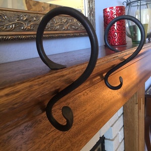 May include: Two black wrought iron decorative fireplace mantel candle holders. The holders have a curved, scroll-like design. A red candle holder and a glass candle holder are in the background. The mantel is made of wood.
