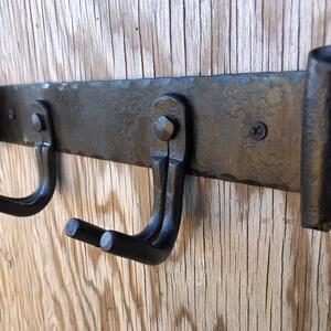 K&ouml;nnte beinhalten: Ein schmiedeeiserner, an der Wand montierter Hakenhalter mit zwei gebogenen Haken. Der Halter ist aus dunklem, geh&auml;mmertem Metall mit dekorativen gerollten Enden. Die Haken sind mit Nieten und Schrauben befestigt, montiert auf einer Holzoberfl&auml;che.