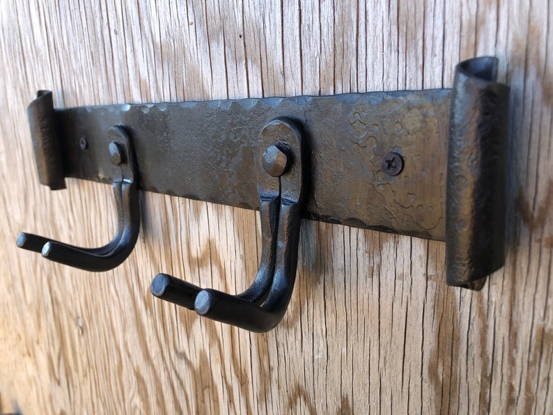 K&ouml;nnte beinhalten: Ein schmiedeeiserner, an der Wand montierter Hakenhalter mit zwei gebogenen Haken. Der Halter ist aus dunklem, geh&auml;mmertem Metall mit dekorativen gerollten Enden. Die Haken sind mit Nieten und Schrauben befestigt, montiert auf einer Holzoberfl&auml;che.