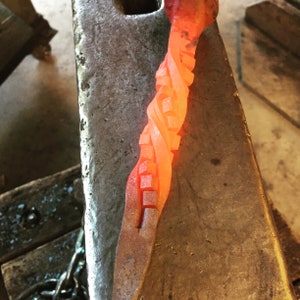 Image of stair-step hot from the forge, appears like a rubik's cube, but is red-hot from being heated in the forge. Laying on anvil.