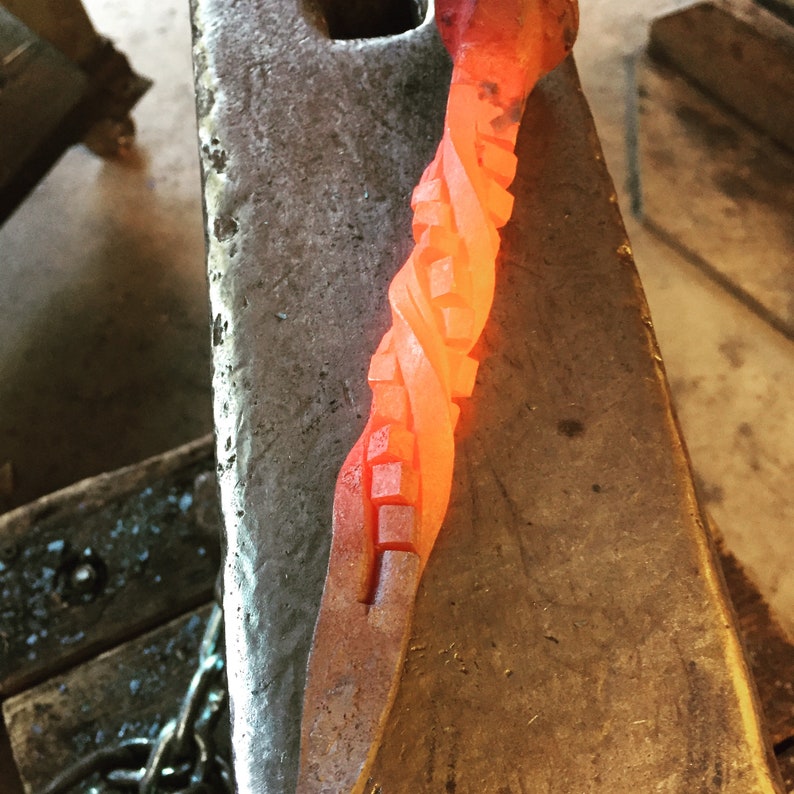 Image of stair-step hot from the forge, appears like a rubik's cube, but is red-hot from being heated in the forge. Laying on anvil.