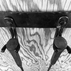 K&ouml;nnte beinhalten: Schwarz-Wei&szlig;-Bild von zwei Metallscharnieren, die an einer Holzoberfl&auml;che befestigt sind. Die Scharniere sind dunkel, mit einem gebogenen Design und einem kreisf&ouml;rmigen Ende. Das Holz hat ein nat&uuml;rliches Maserungsmuster.