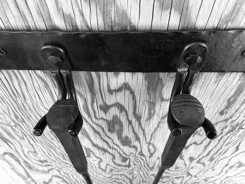 K&ouml;nnte beinhalten: Schwarz-Wei&szlig;-Bild von zwei Metallscharnieren, die an einer Holzoberfl&auml;che befestigt sind. Die Scharniere sind dunkel, mit einem gebogenen Design und einem kreisf&ouml;rmigen Ende. Das Holz hat ein nat&uuml;rliches Maserungsmuster.