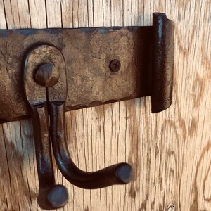 K&ouml;nnte beinhalten: Ein rustikaler, handgeschmiedeter Metall-Kleiderhaken mit zwei gebogenen Haken und einem geraden Haken. Der Haken ist auf einer Holzoberfl&auml;che montiert, die die Textur des Metalls und die Holzmaserung zeigt.