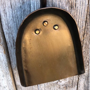 K&ouml;nnte beinhalten: Eine dunkle bronzefarbene Metallschaufel mit einer abgerundeten Schaufel und einem langen Griff. Die Schaufel hat drei kleine, gleichm&auml;&szlig;ig verteilte L&ouml;cher in der N&auml;he der Oberseite. Die Schaufel wird wahrscheinlich zum Umgang mit Asche oder anderen Materialien verwendet.