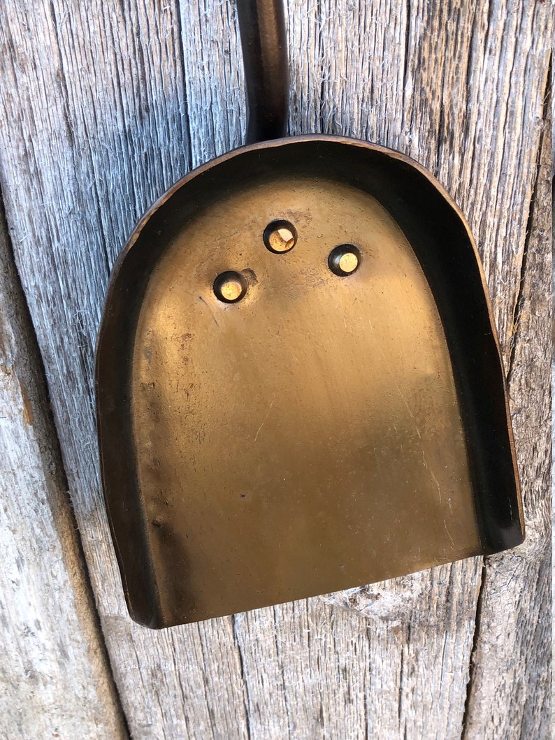 K&ouml;nnte beinhalten: Eine dunkle bronzefarbene Metallschaufel mit einer abgerundeten Schaufel und einem langen Griff. Die Schaufel hat drei kleine, gleichm&auml;&szlig;ig verteilte L&ouml;cher in der N&auml;he der Oberseite. Die Schaufel wird wahrscheinlich zum Umgang mit Asche oder anderen Materialien verwendet.