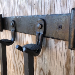 K&ouml;nnte beinhalten: Ein schwarzer, handgeschmiedeter Wand-Kleiderhaken aus Metall mit zwei Haken. Der Haken hat eine strukturierte Oberfl&auml;che und ist auf einem Holzgrund befestigt. Die Haken sind gebogen und zum Halten von Gegenst&auml;nden konzipiert.