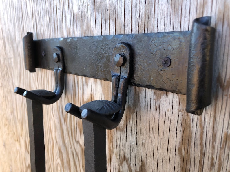 K&ouml;nnte beinhalten: Ein schwarzer, handgeschmiedeter Wand-Kleiderhaken aus Metall mit zwei Haken. Der Haken hat eine strukturierte Oberfl&auml;che und ist auf einem Holzgrund befestigt. Die Haken sind gebogen und zum Halten von Gegenst&auml;nden konzipiert.