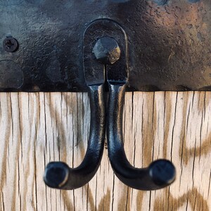 K&ouml;nnte beinhalten: Schwarzer Doppel-Haken-Wandhaken. Der Haken ist aus Schmiedeeisen gefertigt und auf einer schwarzen Metallplatte montiert. Der Hintergrund ist verwittertes, helles Holz.