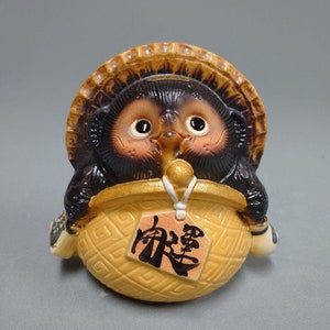 May include: A ceramic Tanuki figurine featuring a dark brown raccoon dog with large eyes, wearing a tan woven hat and holding a light yellow basket with a geometric pattern. A small tag with black Japanese calligraphy hangs from the basket.