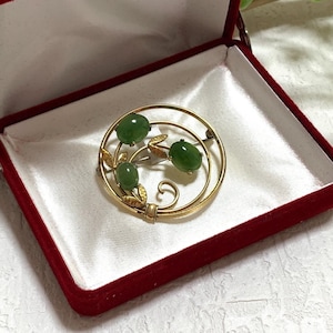 May include: A gold-toned brooch with three green stone accents, presented in an open red velvet jewelry box with a white satin interior. The brooch has a circular design with a floral motif. Greenery is visible in the background.