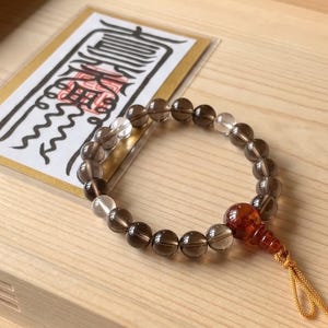 May include: A beaded bracelet featuring translucent smoky quartz spheres, clear quartz accents, and a single reddish-brown gourd-shaped bead with a yellow cord tassel. A rectangular card with black Japanese calligraphy is positioned behind the bracelet.