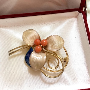 May include: A vintage floral brooch with a gold-tone metal frame, featuring three cream-colored petals, coral-colored beads, and a blue accent. The brooch is presented in a red velvet jewelry box.