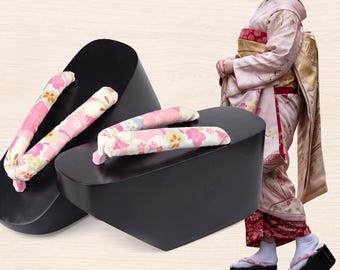 May include: A pair of black wooden platform sandals with thick, elevated soles and fabric straps featuring a pink, white, and yellow floral pattern. The sandals are shown alongside a person wearing a light pink kimono and matching footwear.