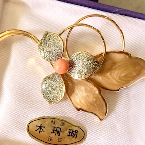 May include: A gold-toned floral brooch with a coral-colored center and glittery accents on the petals. The brooch is in a purple box with a white satin lining. A Japanese label is visible.