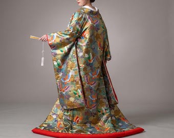 May include: A floor-length gold and multi-colored kimono featuring a repeating pattern of butterflies and floral motifs. The garment has wide sleeves, a red hemline, and is paired with a small handheld fan featuring a white tassel.