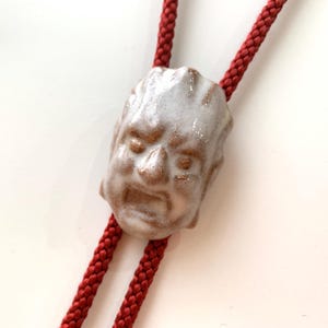 Puede incluir: Una corbata bolo de cerámica con un colgante de rostro esculpido. El colgante tiene un acabado envejecido y vidriado, y está unido a un cordón trenzado rojo. La cara tiene una expresión severa y es blanquecina y marrón.