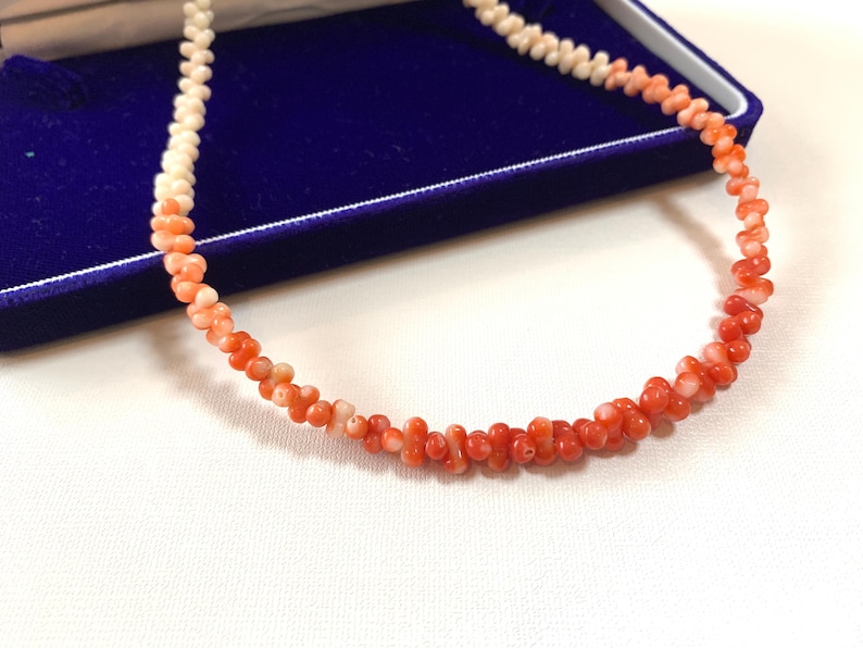 K&ouml;nnte beinhalten: Eine Halskette aus kleinen, unregelm&auml;&szlig;ig geformten, organischen Perlen, die farblich von einem blassen Cremewei&szlig; zu einem leuchtenden Orangerot verlaufen. Die Perlen sind auf einem durchgehenden Strang aufgereiht und erzeugen einen Farbverlauf.