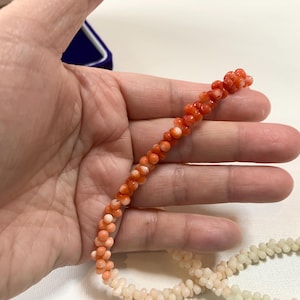 K&ouml;nnte beinhalten: Eine Kette aus kleinen, runden, orangeroten Korallenperlen, die in einem gedrehten, strukturierten Muster angeordnet sind. Die Perlen zeigen einen Farbverlauf von einem leuchtenden Orangerot an einem Ende bis zu einem hellen Cremeton am anderen Ende.
