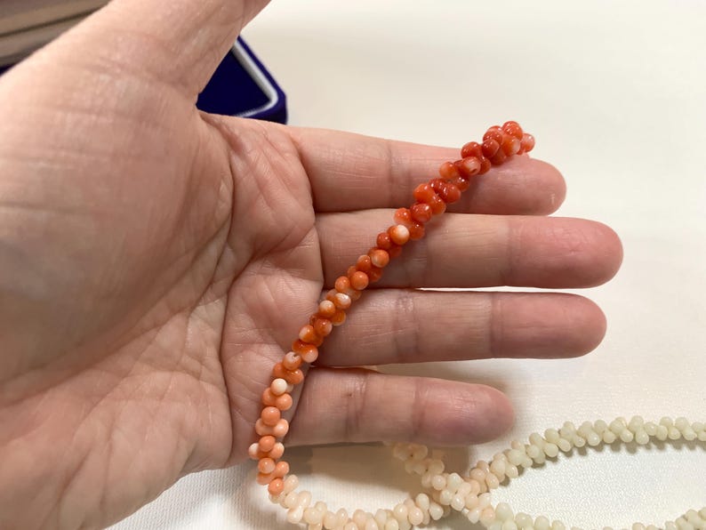K&ouml;nnte beinhalten: Eine Kette aus kleinen, runden, orangeroten Korallenperlen, die in einem gedrehten, strukturierten Muster angeordnet sind. Die Perlen zeigen einen Farbverlauf von einem leuchtenden Orangerot an einem Ende bis zu einem hellen Cremeton am anderen Ende.