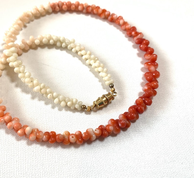 K&ouml;nnte beinhalten: Eine Halskette aus kleinen, texturierten, organisch geformten Perlen in einem Farbverlauf von Cremewei&szlig; bis hin zu leuchtendem Orangerot. Die Kette verf&uuml;gt &uuml;ber einen goldfarbenen, zylindrischen Tonnenverschluss, der die beiden Enden sicher verbindet.