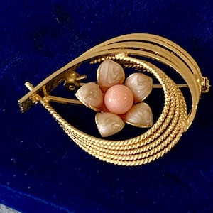 May include: Gold-toned brooch with a floral design. The brooch features a central pink bead surrounded by six petal-shaped elements in cream and white. The brooch has a curved, layered design.