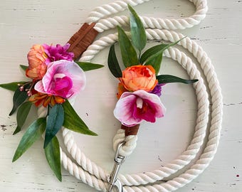 May include: White rope dog lead with a floral accent. The lead has a silver clip and is decorated with pink, orange, and green flowers.