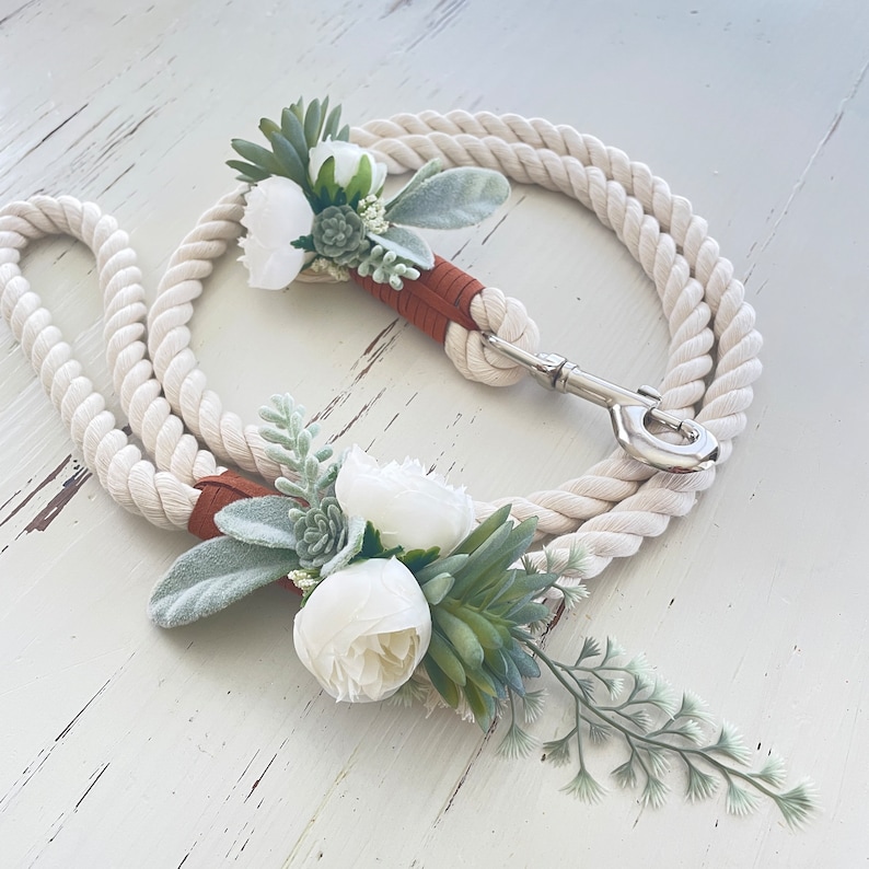 May include: White rope dog leash with a brown leather handle and two white flower attachments. The flowers are made of fabric and have green leaves.