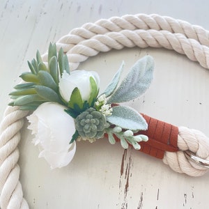 May include: A white and green floral arrangement with a brown leather strap attached to a white rope. The flowers are arranged in a cluster and include white roses, green succulents, and white sprigs.