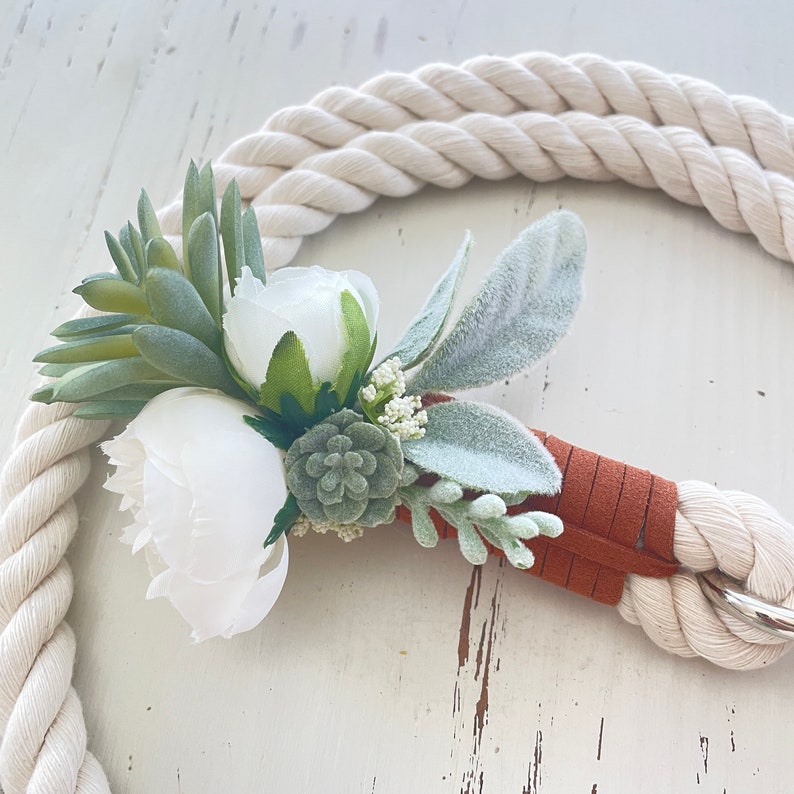 May include: A white and green floral arrangement with a brown leather strap attached to a white rope. The flowers are arranged in a cluster and include white roses, green succulents, and white sprigs.