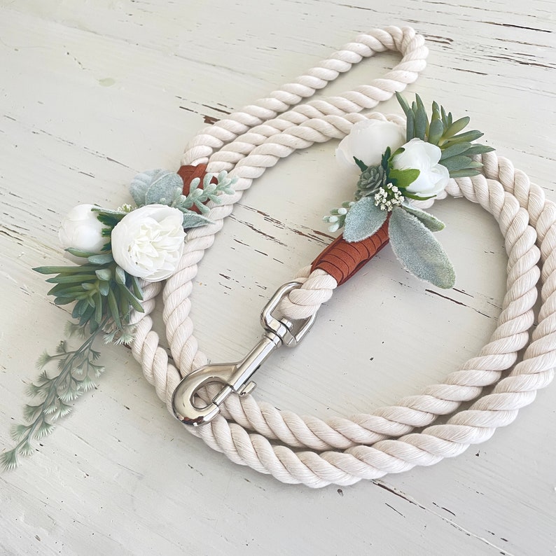May include: A white rope dog leash with a brown leather handle and white flower accents. The leash has a silver metal clasp.