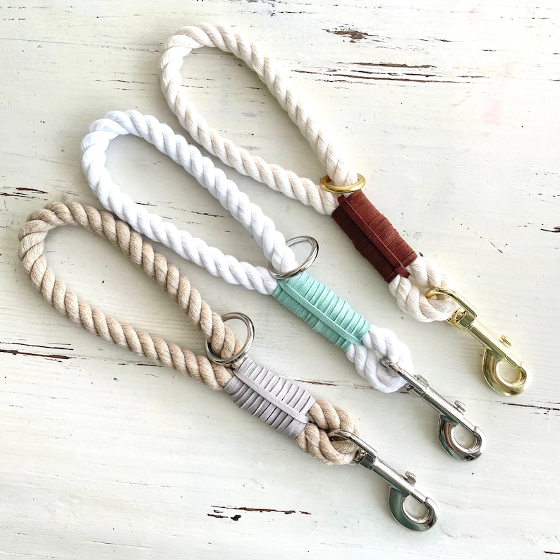 May include: Three dog leads with thick, twisted rope handles. One lead is off-white with a brown leather accent and gold-coloured clasp. The other two are white and tan with silver clasps and mint and grey accents.