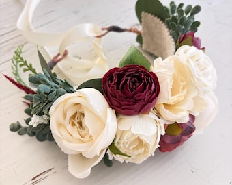 Pode incluir: Uma coroa de flores com rosas em tons de creme e bordô escuro, acentuada com folhagem verde e pequenos ramos brancos. O arranjo está fixado numa base fina e flexível com longas fitas de cetim cor de creme que se estendem para trás.
